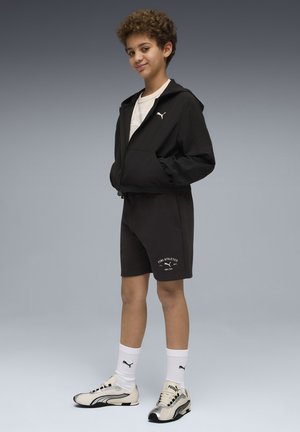 Garçon debout, les mains dans les poches d'un sweat à capuche noir, portant un short Puma noir, des chaussettes blanches Puma et des baskets blanches et noires, sur un fond gris.