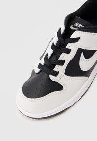Crni i bijeli Nike tenisica s crnim vezicama, perforiranim prednjim dijelom i Swoosh logotipom sa strane, prikazana na jednostavnom svijetlom pozadini.