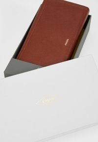 Portefeuille en cuir marron dans une boîte grise, arborant un logo doré "FOSSIL". Le portefeuille présente une finition texturée avec des coutures soignées.