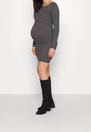 Femme enceinte debout, portant une robe moulante en maille côtelée grise et des bottes noires montantes jusqu'aux genoux sur un fond blanc uni.