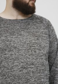 Grå strikkegenser med et teksturert, melert mønster og vid halslinning. Har raglan-ermer og myk stoff.