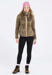 Fuzzy bruine zip-up jas met hoge kraag, contrasterende tan rits en voorzakken. Gecombineerd met zwarte leggings en bruine wandelschoenen.