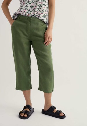 Osoba nosiaca zelené krátke nohavice, kvetinový tielko bez rukávov a čierne sandále so sponou, stojaca s rukami vreckách.