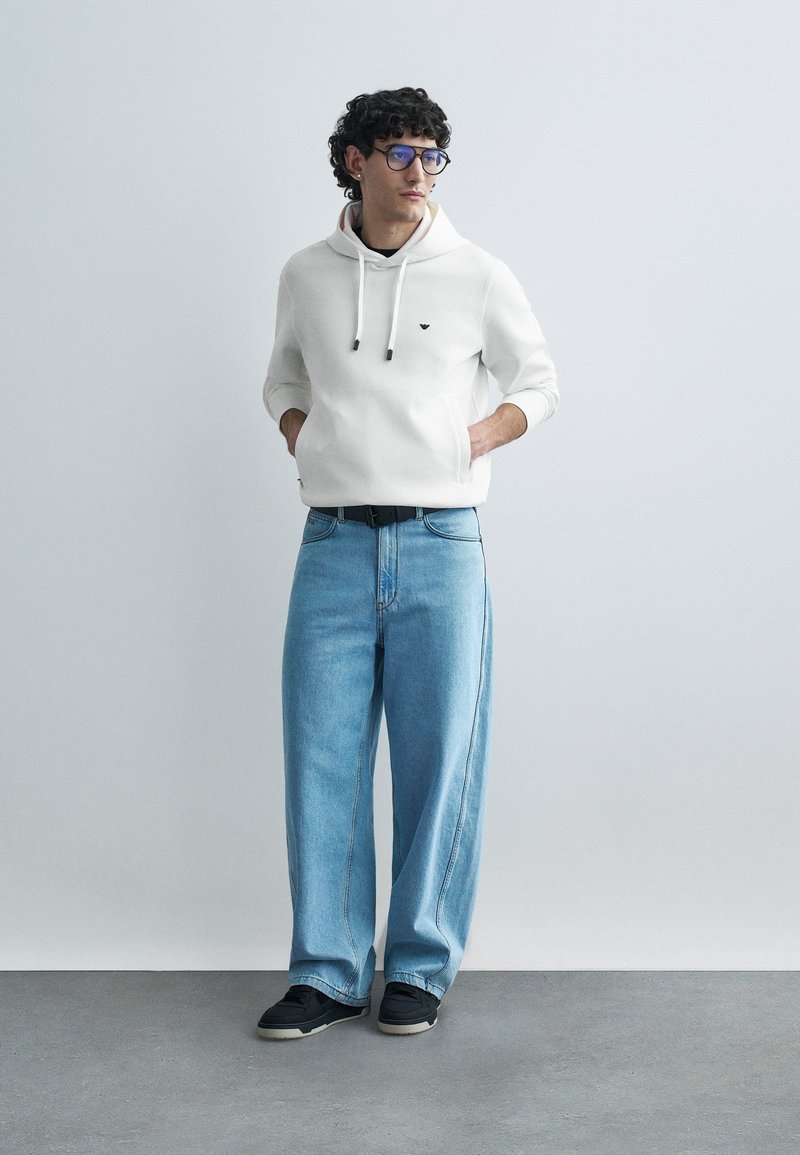 Balta džemperio su gobtuvu, su raišteliais, juodu logotipu, derinama su šviesiai mėlynais plačiomis kojomis džinsais ir juodais sportbačiais. Fonas neutrali ir minimalistinė.