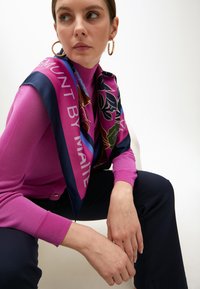 Seidenschal mit floralem Muster in Lila, Marineblau und Grün, über einem rosa Strickpullover mit gerippten Bündchen drapiert. Modell in dunklen Hosen, sitzt.