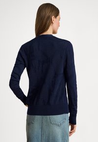 Tmavomodrý pletený kardigán so štruktúrovaným vzorom, rebrovanými manžetami a spodným okrajom, a klasickým okrúhlym výstrihom, nosený cez svetlú denimovú sukňu.