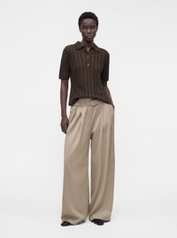 Personne debout, face à l'avant, portant une chemise marron à manches courtes avec col et un pantalon beige taille haute à jambes larges, ceinturé, mains dans les poches.