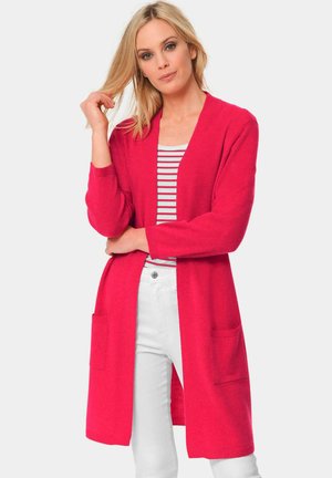 Cardigan long, à l'avant ouvert, en tissu tricoté rose vif avec des manches longues et deux poches à l'avant, porté sur un haut rayé et un pantalon blanc.