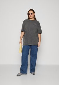 Camiseta gris de gran tamaño con texto, combinada con jeans rectos azules. Lleva gafas de sol geométricas y sostiene un pequeño bolso amarillo.