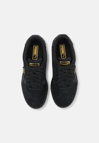 Černé semišové tenisky s texturovaným povrchem, zkombinované s černými tkaničkami, zlatým logem a gumovou podrážkou. Bez viditelných vzorů nebo akcentů.