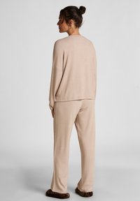 Beige loungewear-set gemaakt van zachte stof, met een losse fit lange mouw top en bijpassende losse broek, aangevuld met donzige donkerbruine slippers.