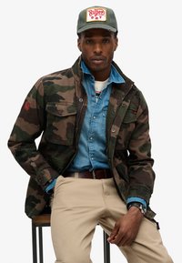 Camo jack, denim overhemd met knoopdetails, beige broeken, bruin riem, grijze pet met rood logo patch, gestructureerde stof, comfortabele pasvorm.