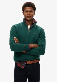 Sudadera de polar verde con cremallera y cuello alto, superpuesta sobre una camisa de cuadros en tonos rojos y verdes. Jeans oscuros y un cinturón marrón completan el look.