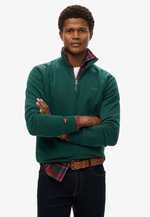 Pullover in pile verde con zip e colletto alto, indossato sopra una camicia a quadri con tonalità di rosso e verde. Jeans scuri e una cintura marrone completano il look.