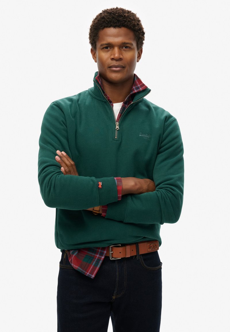 Sudadera de polar verde con cremallera y cuello alto, superpuesta sobre una camisa de cuadros en tonos rojos y verdes. Jeans oscuros y un cinturón marrón completan el look.