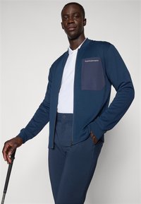 Marineblaue Zip-Jacke mit einer dunkleren Brusttasche, kombiniert mit passenden, enganliegenden Hosen. Mit langen Ärmeln und einem klassischen Kragen.
