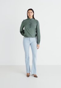 Blusa verde con texture e colletto a volant, abbinata a jeans svasati azzurri. Modella che indossa tacchi con stampa animale, in piedi su uno sfondo bianco.