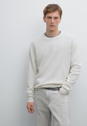 Maglione a maglia color crema con maniche lunghe abbinato a pantaloni grigi chiari. La texture sembra morbida, con scollatura rotonda e dettagli sottili.