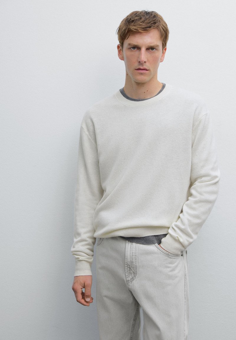 Crèmefarbener Strickpullover mit langen Ärmeln, kombiniert mit hellgrauren Hosen. Die Textur wirkt weich, hat einen runden Ausschnitt und dezente Details.