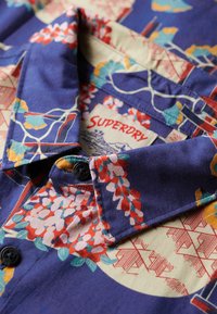 Korte mouwen overhemd van blauwe stof met kleurrijke bloem- en geometrische patronen. Voorzien van een kraag en zwarte knopen; label zichtbaar.
