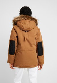 Brauner Kapuzenparka mit Kunstpelzbesatz, schwarzen Ellbogenflicken und tailliertem Schnitt. Strukturierter Stoff und lockere Passform. Rückansicht.