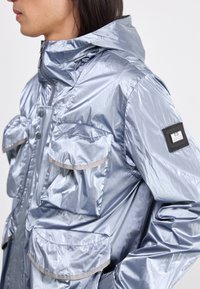 Light blue metallic rain jacket with a hood, featuring multiple front pockets, gray accents, and a logo patch on the left sleeve.