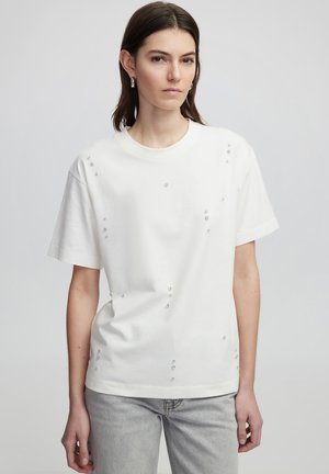 T-shirt en coton blanc à manches courtes, ornée de broderies en forme de gemmes claires disposées de manière asymétrique sur le devant.
