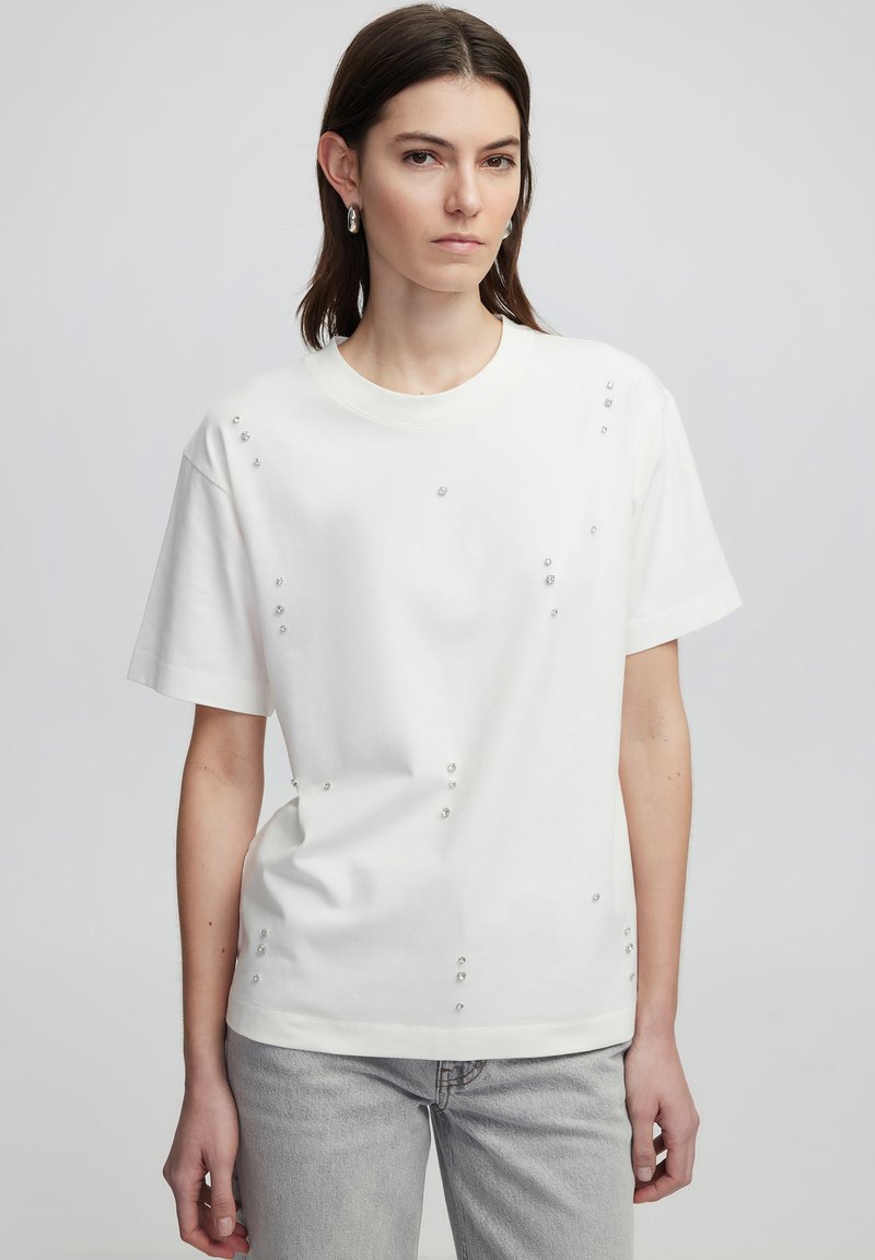T-shirt en coton blanc à manches courtes, ornée de broderies en forme de gemmes claires disposées de manière asymétrique sur le devant.