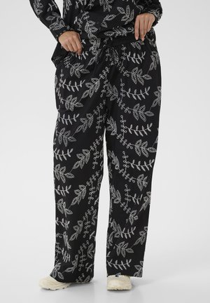 Pantalon noir à jambes larges avec motifs blancs de feuilles et de vignes, porté avec un haut assorti et des baskets blanches, mains ajustant la taille.