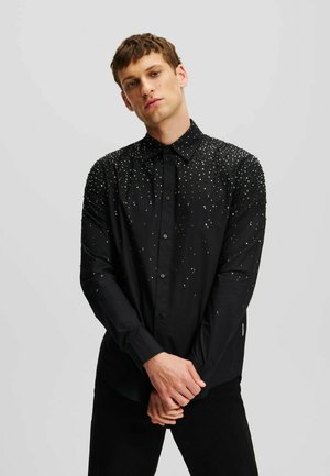 Joven con una camisa negra de manga larga con botones y patrones moteados blancos, de pie con las manos entrelazadas delante.