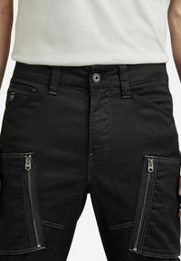 Svarta byxor med dragkedjefickor på båda låren, metallknapstängning, bälteshällor och en liten logopatch på fickan till höger.