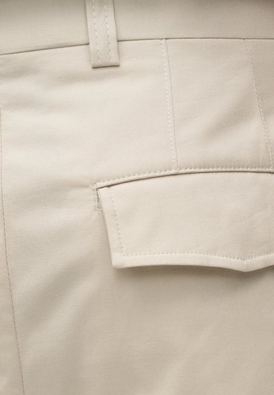 Κοντινή λήψη μπεζ υφάσματος με ραμμένη τσέπη και θηλιά ζώνης, που δείχνει υφή και λεπτομέρειες ραφής.