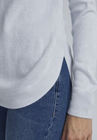 Hvid striktrøje med ribbede manchetter, en let afsmalnet hollænder og sideslids. Kombinationen med mørkeblå denim fremhæver tekstur og pasform.