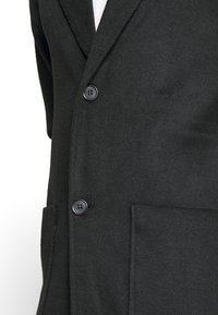 Blazer in lana nera con vestibilità su misura, design monopetto, due bottoni e due tasche frontali. Tessuto morbido e finitura liscia.