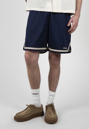 Hombre vistiendo pantalones cortos de malla azul marino con ribete beige, calcetines blancos con logo y zapatos de ante beige con cordones, de pie frente a un fondo liso.