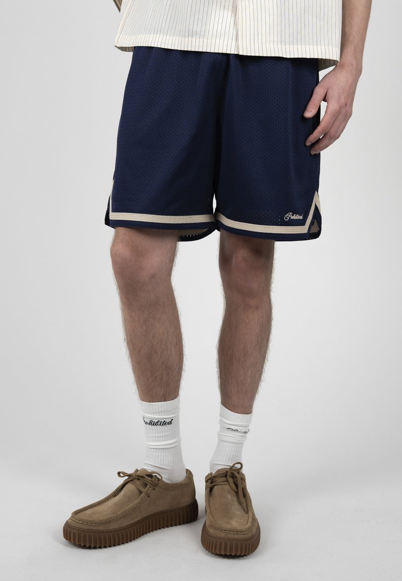 Homme portant un short en mesh bleu marine avec bordure beige, des chaussettes blanches avec logo et des chaussures en suède beige à lacets, debout devant un fond uni.