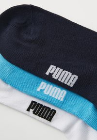Tris Puma kojinių porų: vienos tamsiai mėlynos, vienos šviesiai mėlynos su juodu tekstu ir vienos baltos su juodu tekstu, visos su sklandžiu paviršiumi.