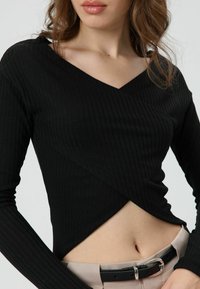 Zwarte geribbelde top met lange mouwen, een V-hals en een overslagontwerp, met een cropped zoom en subtiele textuur. Gecombineerd met beige broek en een zwarte riem.