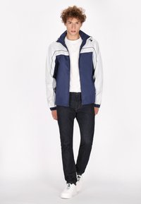 Blauwe en witte windbreaker jas met een ritssluiting aan de voorkant, elastische mouwen en een trekkoord capuchon, gedragen met donkere jeans en witte sneakers.