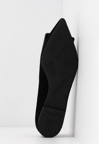 Chaussure plate noire pour femme à bout pointu reposant sur le côté, semelle montrant un logo embossé et des motifs décoratifs de chaussons de ballet.