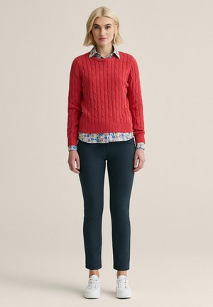 Kvinde med kort blondt hår iført rød kabelstrikket sweater over blomstret skjorte, mørke smalle bukser og hvide sneakers, stående mod ensfarvet baggrund.