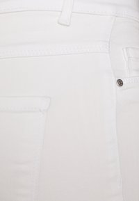 Jean de mezclilla blanca con diseño de cinco bolsillos, costuras sutiles y un botón de logo plateado cerca del bolsillo derecho.