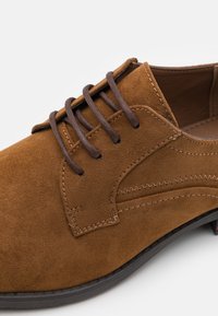 Pantof brown din suede, cu șireturi, cu o textură fină, vârf rotund, cusături contrastante și ochiuri de șireturi întunecate. Talpa este neagră și ușor texturată.