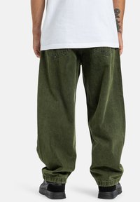 Zielone dżinsy typu wide-leg z teksturowym wykończeniem, z tylnymi kieszeniami i mankietami na elastykach. Noszone z jednolitą białą koszulką i czarnymi butami.