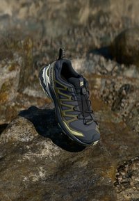 Black and olive green athletic shoe with a mesh upper, textured material, distinctive lacing system, and rugged sole for traction.