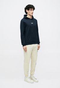 Felpa con cappuccio blu navy con coulisse e tasca frontale, abbinata a pantaloni della tuta color crema. Il modello indossa sneakers chiare. Design semplice e casual.