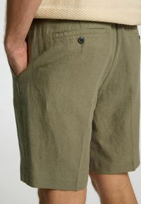 Olivza zaļas linu šortus ar sānu kabatām, pogu aizdari un nedaudz strukturētu apdari. Apakša atrodas virs ceļa, nodrošinot brīvu piegulšanu.