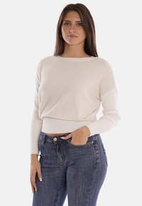 Maglione cropped color crema con un corpo testurizzato e maniche a costine. Abbinato a jeans attillati blu scuro, che mostrano un look casual e contemporaneo.