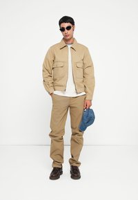 Beige bomber jacket with two front pockets, white t-shirt, light brown pants, and brown shoes. Holding a blue baseball cap.