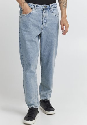 Mand iført lyseblå løstsiddende jeans og sorte sneakers med hvide såler, stående mod en ensfarvet lys baggrund.